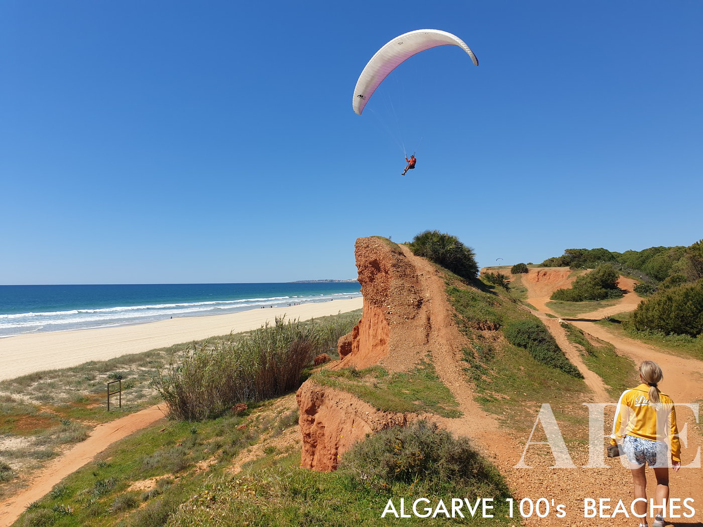 Vue ouest en mars. Ambiance sereine sur la plage Falésia de Rocha Baixinha. Avec un sentier pédestre à flanc de falaise qui en trace le contour, la plage devient une scène d'activité tranquille. Les parapentes parsemant le ciel clair ajoutent une dimension exaltante, tandis que les amateurs de plage, profitant d'une promenade sur le sable immaculé, contribuent à l'atmosphère vibrante mais paisible