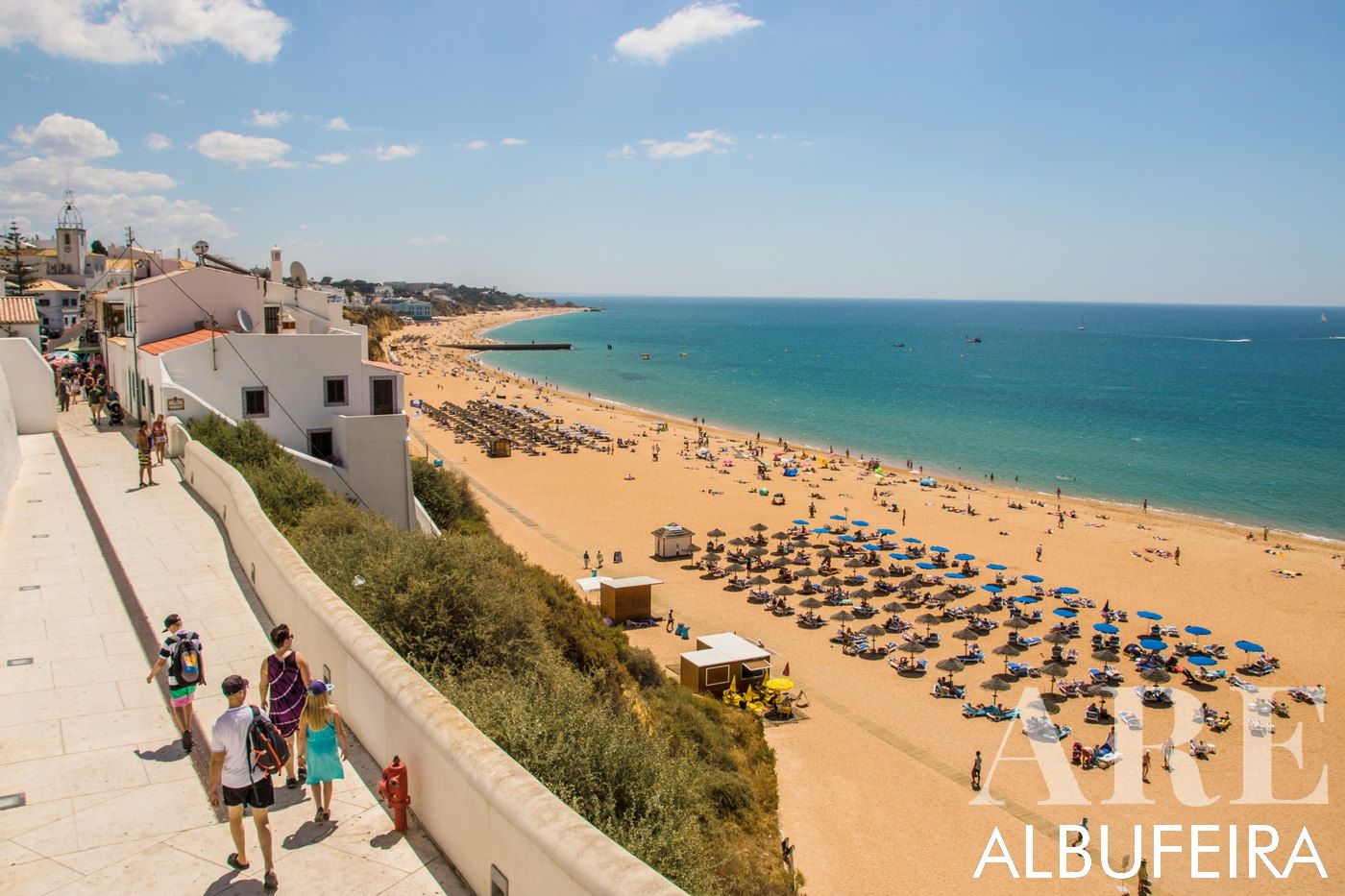 Eastern view of Albufeira's beach and the old town. On the left, a family strolls on the elevated pathway leading to the old town, with quaint cliffside houses beneath. Below, the sandy beach is speckled with umbrellas, bordered by the greenish hues of the ocean, all under a clear, blue sky