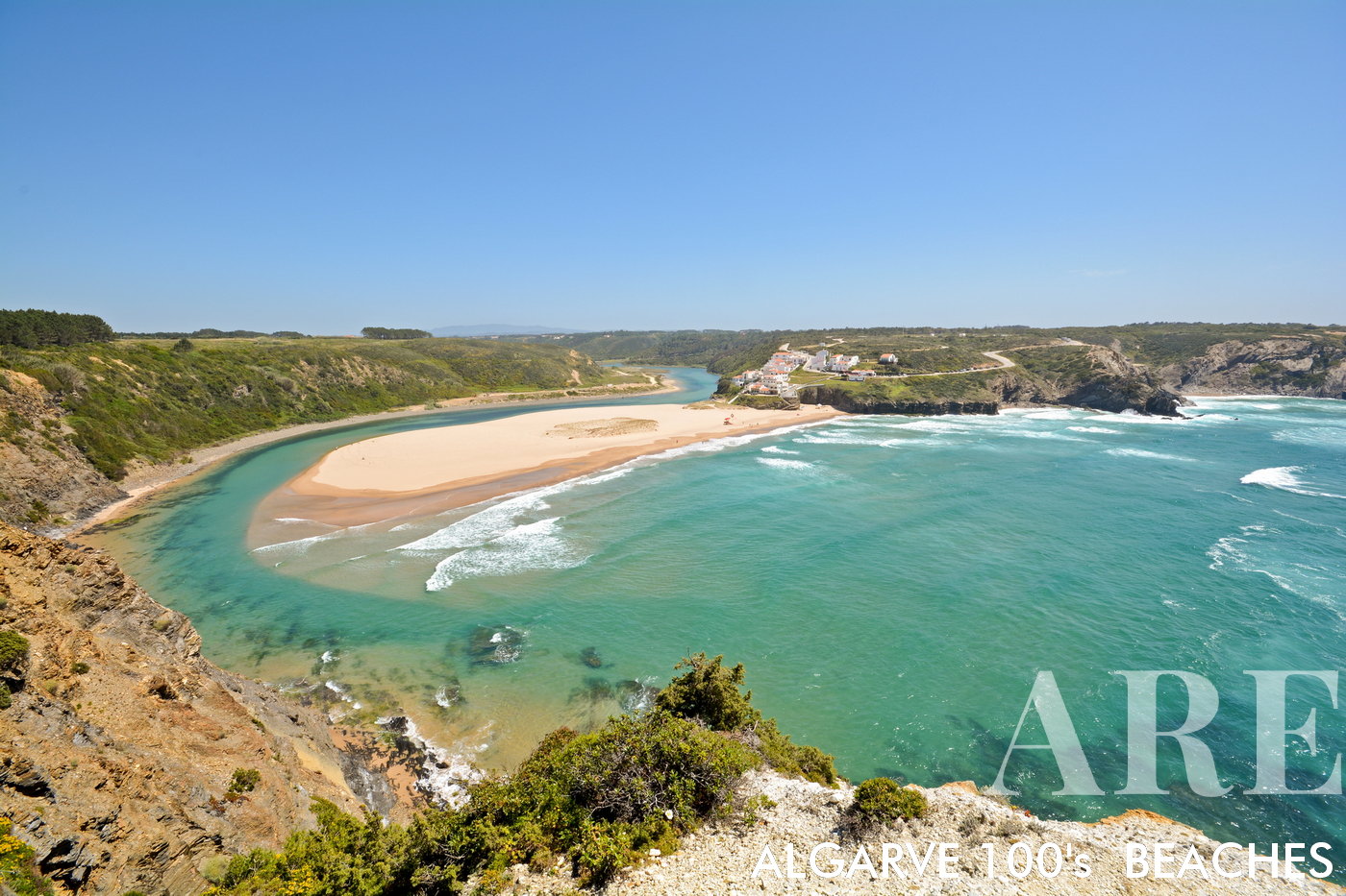 A panoramic view opens up to Odeceixe Beach, a surfer's paradise on the West coast of Algarve, in the Aljezur District of Portugal. A sandy bank, shaped by the river, graces the scene, while the river meets the ocean on the right side of the beach. On the left, a small village of quaint houses perches on the cliff top, overlooking the scenic coastal vista.