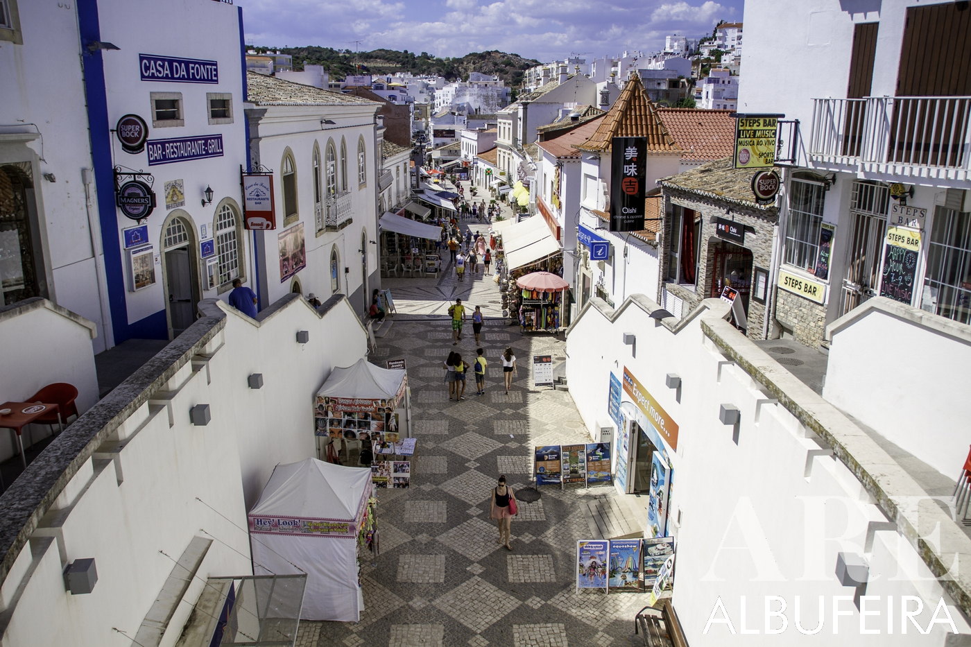 Albufeira city center street, paved with traditional 'pedra de calçada', bustling with a variety of shops offering souvenirs, lively bars, diverse restaurants, and an array of liquors.