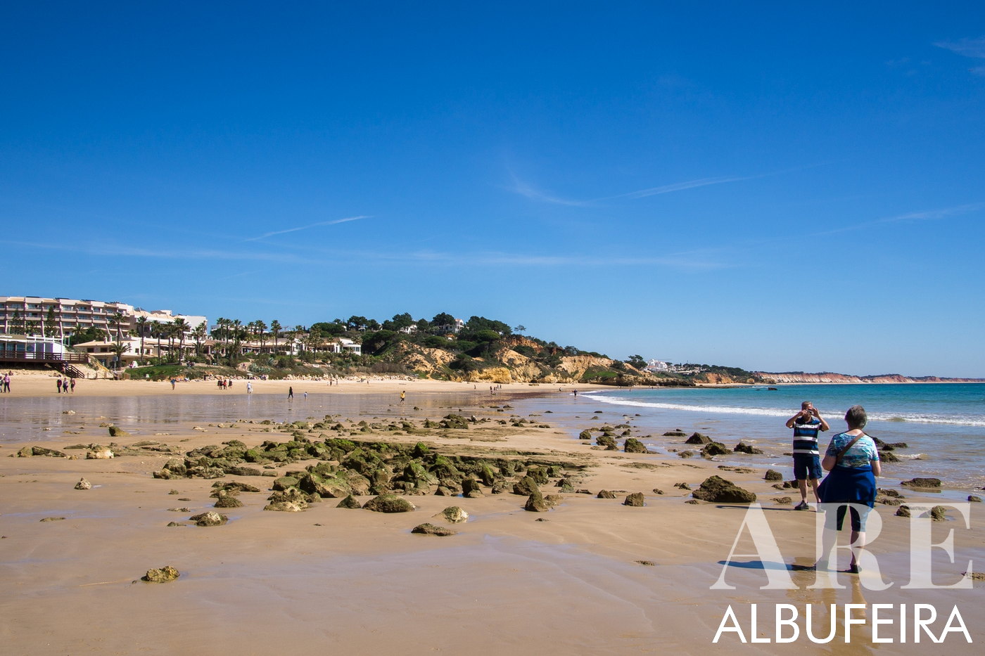 view of Santa Eulalia Beach with the distant Praia da Falésia visible on the horizon. Low tide reveals striking green rocks adorned with limes, contrasting against the lengthy stretch of sandy beach.
