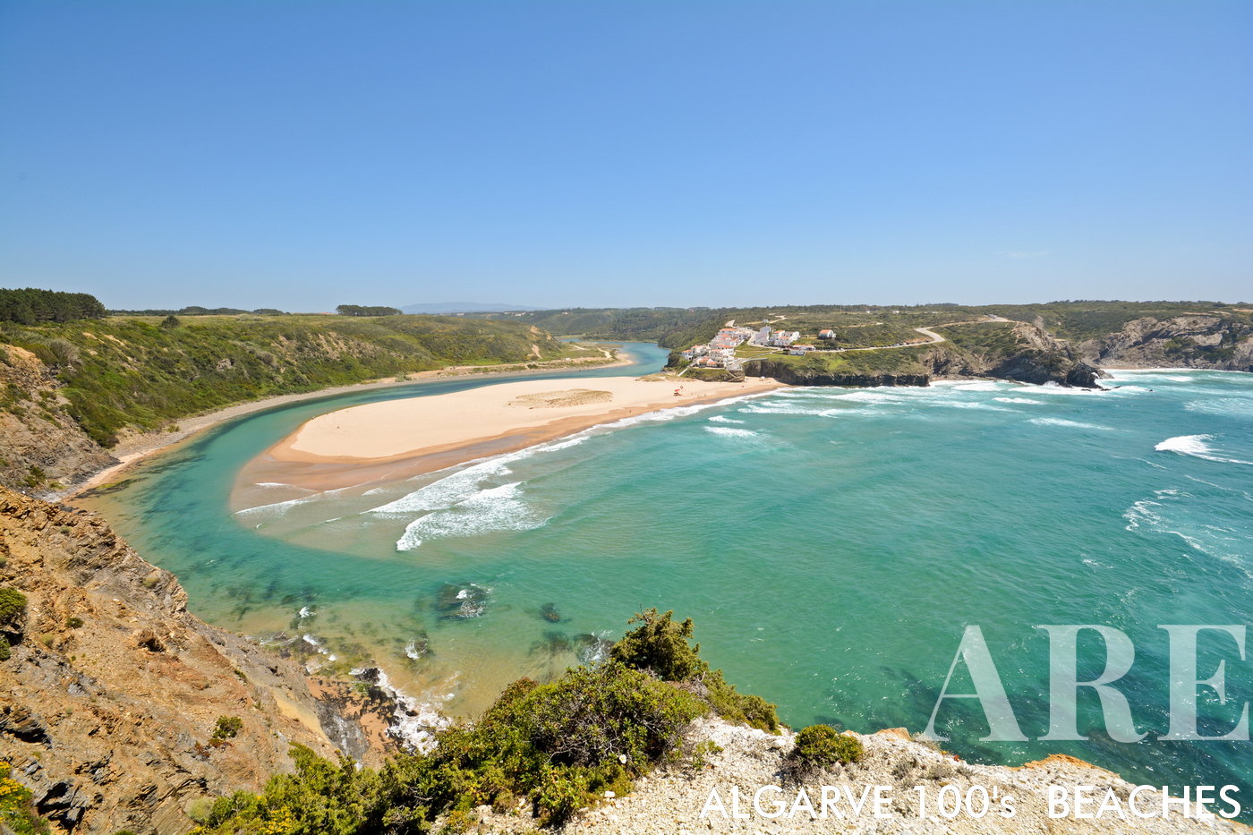 Uma vista panorâmica abre-se para a Praia de Odeceixe, um paraíso para os surfistas na costa oeste do Algarve, no distrito de Aljezur, em Portugal. Um banco de areia, moldado pelo rio, enfeita a cena, enquanto o rio encontra o oceano do lado direito da praia. À esquerda, uma pequena aldeia de casas pitorescas empoleira-se no topo da falésia, com vista panorâmica para a costa.