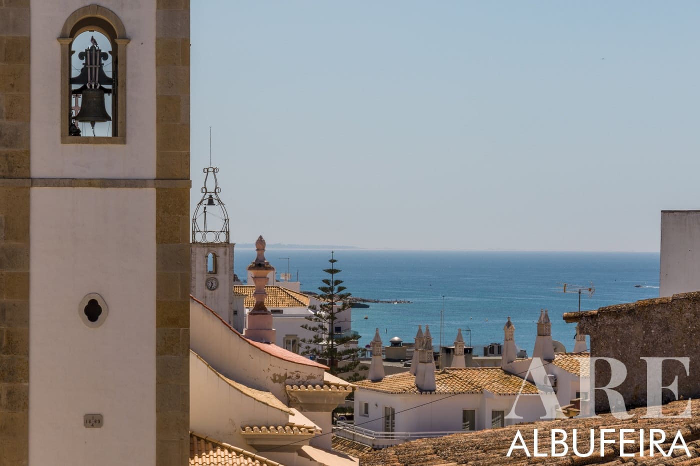 O sino e a torre da igreja de Albufeira erguem-se majestosamente acima de um pitoresco horizonte de casas, tudo tendo como pano de fundo o vasto oceano no horizonte.