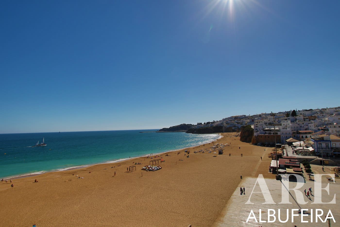 Espectacular miradouro com vista para a Praia dos Pescadores de Albufeira e para a encantadora Zona Velha à direita. O oceano azul se espalha à direita, enquanto a praia de areia se aninha centralmente abaixo sob um céu azul amplo e claro.