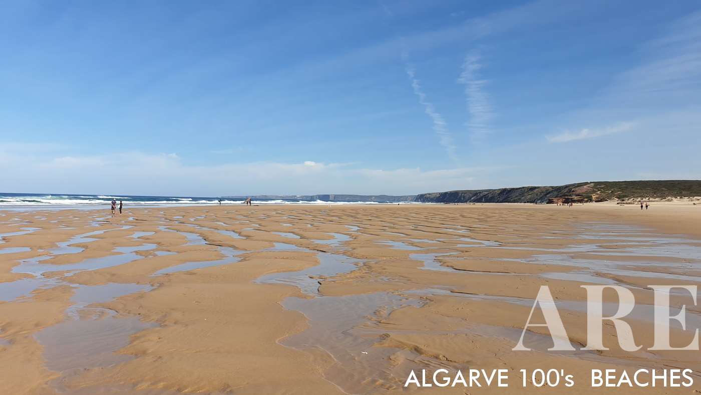 Bordeira Beach features a vast sandy area, accentuated by sand banks revealed at low tide. The beach is bordered by cliffs lush with green vegetation.