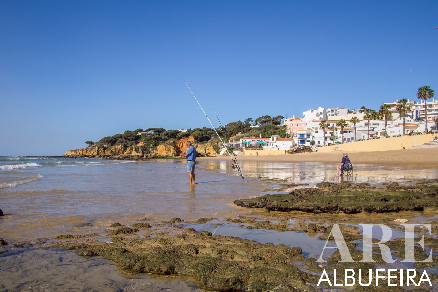 February 23rd scene at Praia de Olhos d ' Água, Albufeira. Features include fishermen with cane poles, low tide revealing water-logged rocks, marking the eastern village limit of Albufeira. To the further east lies Açoteias, a quaint village nestled just beyond the beachfront.