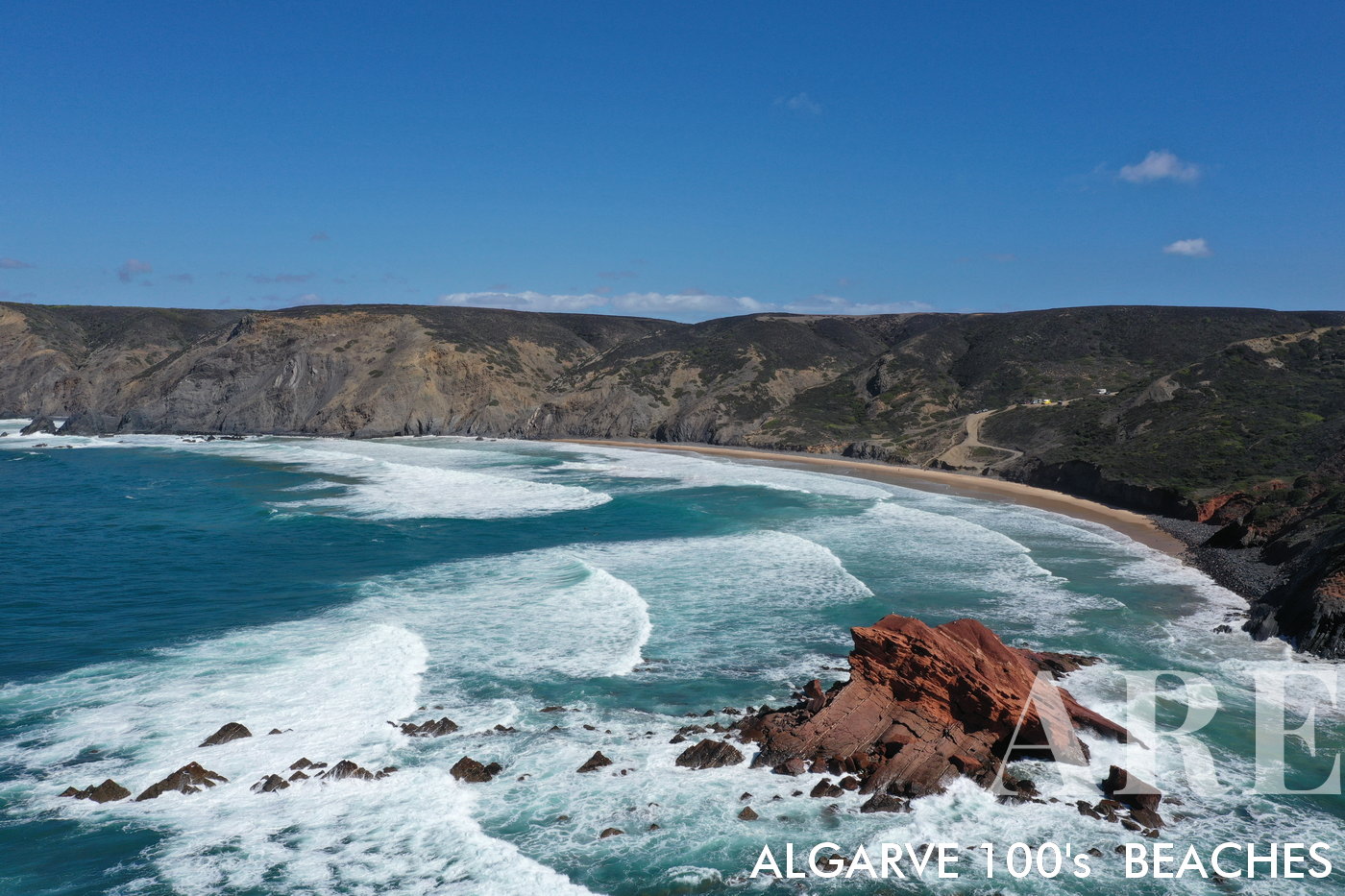 Vista cativante da Praia de Ponta Ruiva a partir do oceano, destacando-se a famosa rocha Ruiva à direita, as ondas dançando da esquerda para a direita e uma dramática falésia enfeitando o lado direito da imagem. Um paraíso de surfe secreto, acessado apenas por uma estrada de terra isolada.