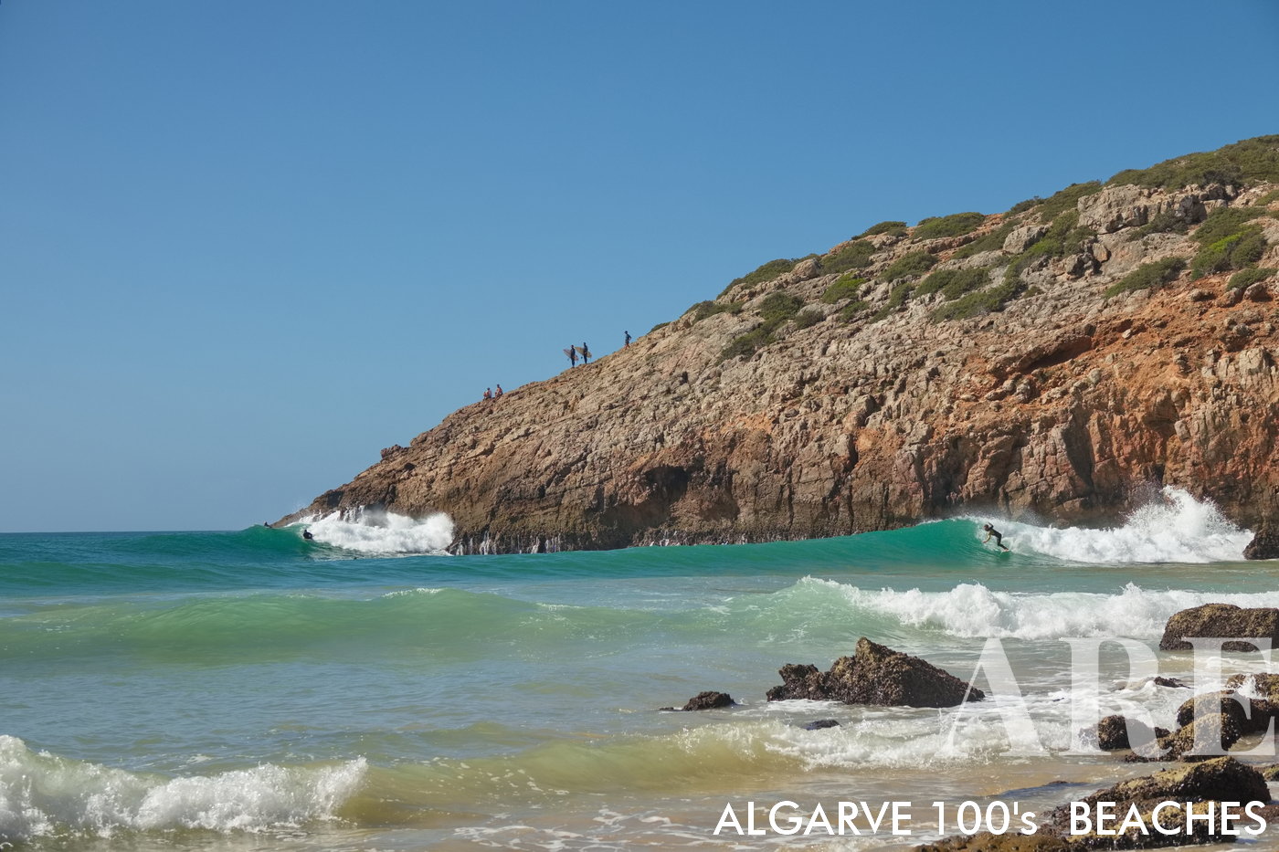 At Zavial Beach, the western waves crash dramatically, amplified by the nearby cliff. Silhouettes of surfers dot the cliff top, while others ride the waves below, creating a dynamic tableau of adventure and nature
